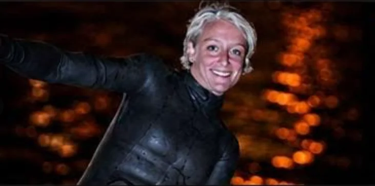 Sara Campbell smiling in a wetsuit against dark water with orange light reflections